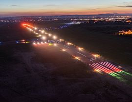 Widok z lotu ptaka na oświetlony pas startowy krośnieńskiego lotniska o zmierzchu. Wzdłuż drogi startowej świecą światła nawigacyjne, a w tle widać panoramę miasta.